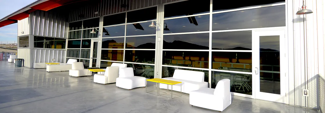 Outdoor patio with sleek white seating and yellow tables against a modern building with large glass windows. Mountains are reflected in the glass. Atmospheric.